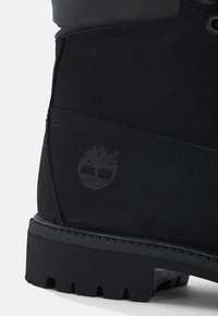 Sección del tacón de una bota negra robusta con costuras visibles y un logo de árbol en relieve en el lateral, mostrando una suela texturizada antideslizante.