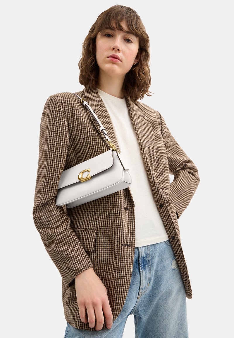 Femme portant un blazer à carreaux marron, chemise blanche, jean bleu clair, portant un sac à main blanc avec un fermoir doré sur l'épaule.