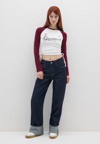 Wit en kastanjebruin bovenstuk met lange mouwen en "Liverpool" print met zijstrikjes, gecombineerd met donkerblauwe wijde jeans en chunky witte sneakers.
