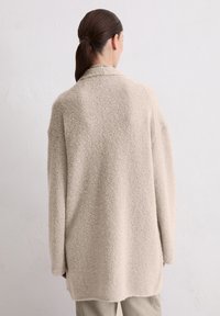 Beige, fuzzy jas met een gestructureerde afwerking, lange mouwen en een ontspannen pasvorm. Heeft een kraag en komt tot halverwege de dij. Achteraanzicht.