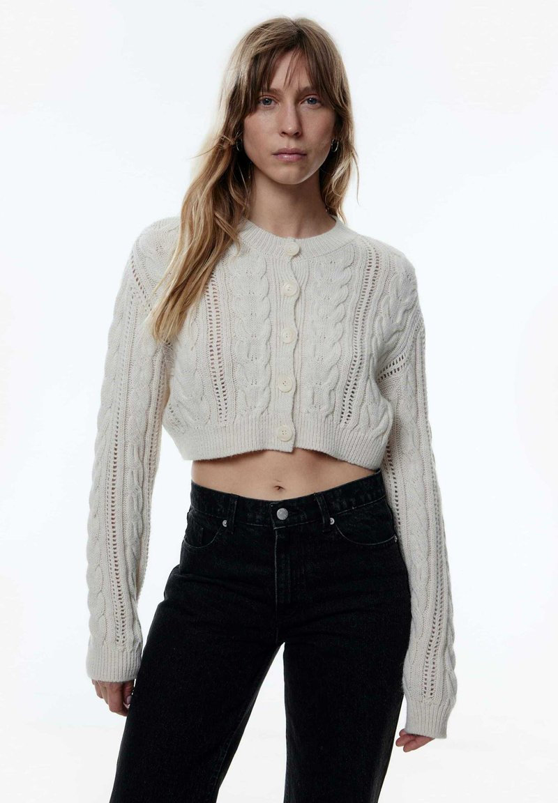 Cardigan créme en tricot épais avec des motifs en câble et des oeillets, design court, encolure ronde, et six boutons à l'avant associé à un jean noir.