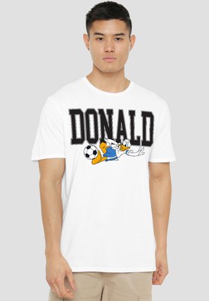 Young man wearing white t-shirt with "DONALD" text and cartoon duck playing soccer on front, beige pants, neutral background.