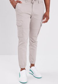 Beige cargo broeken met elastische boorden, voorzien van vier zakken en zichtbare stikseldetails, gedragen met witte sneakers en groene accenten.