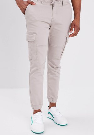 Pantaloni cargo beige con polsini elasticizzati, dotati di quattro tasche e dettagli di cucitura a vista, indossati con sneakers bianche e accenti verdi.