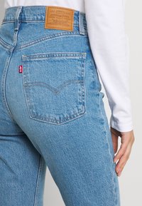 Svetle modré džíny s vysokým pasem, které mají koženou nášivku s nápisem "Levi Strauss & Co." a červenou tabulku na zadní kapse.