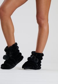 Black plush slippers with a ribbed design, featuring pom-pom accents, soft texture, and a white sole. Suitable for indoor wear.