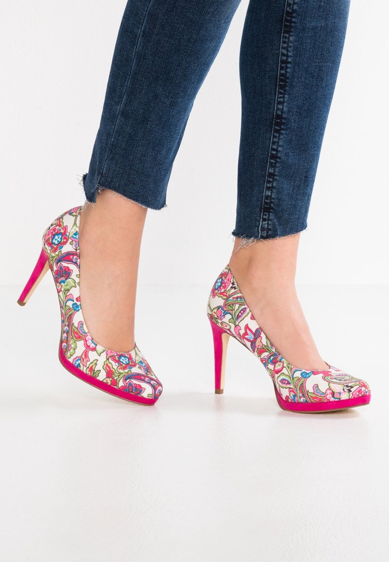 Jambes portant un jean court bleu foncé et des talons hauts à motif floral avec des semelles et des talons roses, debout sur un sol blanc.