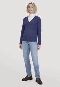V-Ausschnitt-Pullover aus tiefblauer Strickware mit langen Ärmeln, kombiniert mit hellblauen Jeans und dunkelbraunen Schuhen, vor einem einfarbigen Hintergrund stehend.