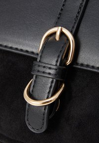Curea pentru geantă din piele neagră și suede, cu o cataramă din metal auriu și detalii de cusătură. Textura contrastează între neted și moale.