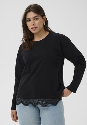 Kvinde med mørkt hår iført sort langærmet top med blondekanter og lyseblå jeans, poserer med den ene hånd på hoften mod en ensfarvet baggrund.