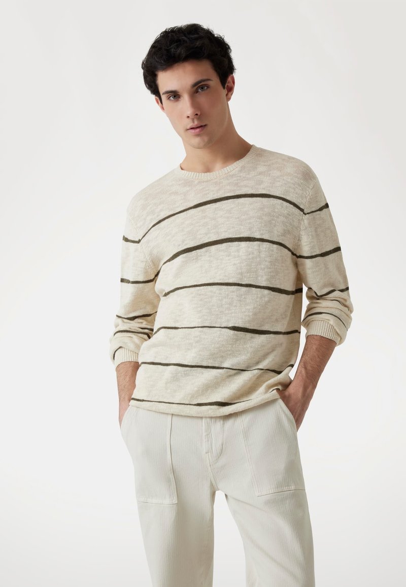 Pull en tricot couleur crème avec des rayures horizontales vert foncé, col ras du cou, poignets côtelés et coupe légèrement ample. Associé à un pantalon de couleur claire.