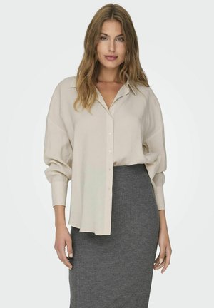 Beige oversized button-up shirt with a collar, featuring long sleeves and a relaxed fit, paired with a gray ribbed pencil skirt.