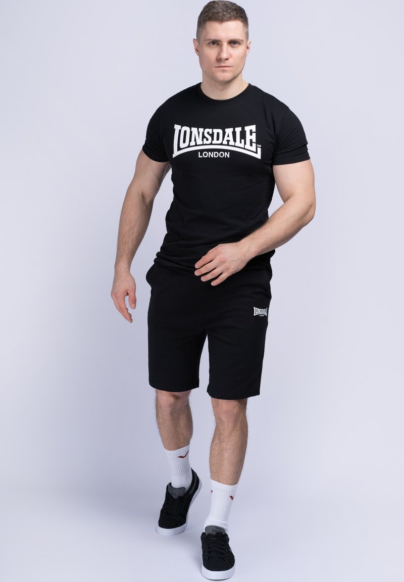 Zwarte katoenen T-shirt met witte "LONSDALE LONDON" logo, gecombineerd met zwarte shorts. Model draagt witte sokken en zwarte schoenen.