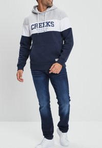 Marineblauer und grauer Hoodie mit einem weißen Streifen, versehen mit dem Text "CREEKS" in Marineblau. Kombiniert mit dunklen Jeans und weißen Sneakers.