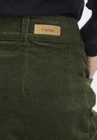 Vihreät corduroypantsit, joissa on pystysuorat raidat, varustettuna ruskealla tarralla, jossa lukee "fransa", sekä sivutaskuilla. Materiaali on pehmeä ja teksturoitu.