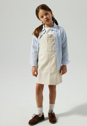 Lichtblau gestreepte shirt over een witte T-shirt, beige denim jurk met zakken, witte geribde sokken en bruine schoenen.