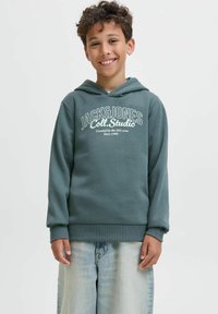 Ragazzo sorridente con capelli ricci che indossa un hoodie teal Jack & Jones e jeans azzurri, in piedi davanti a uno sfondo bianco semplice.