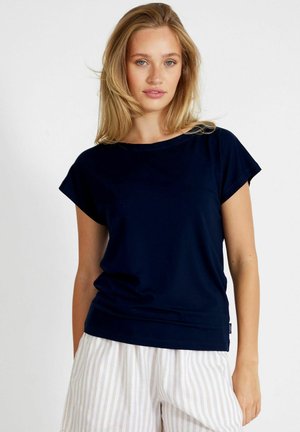 Blondide juustega naine, kes kannab lihtsat tumesinist lühikeste varrukatega särki ja valgete triipudega pükse, valge tausta ees.