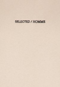 T-shirt in cotone beige con testo ricamato in nero "SELECTED / HOMME" centrato sul petto. Texture liscia con design aderente.