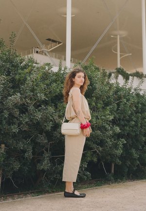 Młoda kobieta o kręconych włosach, ubrana w beżowy strój, czarne buty, czerwoną gumkę do włosów i kremową torebkę, stoi na zewnątrz obok zielonych krzewów.