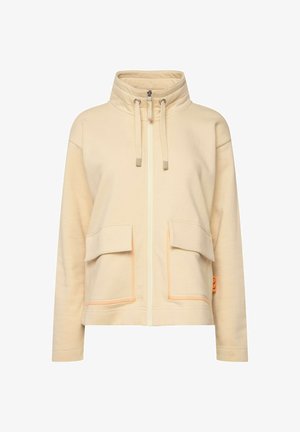 Beige ritsjack met hoge kraag, twee voorkruisen, oranjetinten en zachte stof. Kenmerken zijn de trekkoorden voor een verstelbare pasvorm.