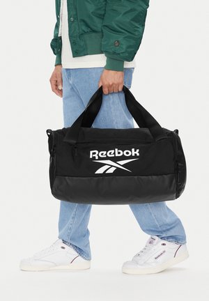 Osoba w zielonej kurtce i niebieskich dżinsach trzyma czarną torbę Reebok z białym logo, ma na sobie białe buty Reebok.