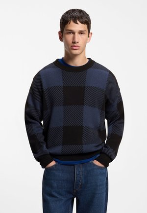 Ung mand med kort mørkt hår iført en sort og blå ternet sweater og blå jeans, stående med hænderne i lommerne mod en hvid baggrund.