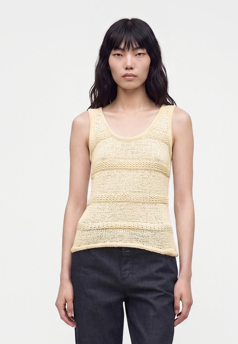 Femme aux longs cheveux noirs portant un haut en tricot crème sans manches et un pantalon foncé, debout devant un fond clair uni.