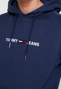 Närbild av en marinblå huvtröja med vit text "TOMMY JEANS" och en liten rektangulär logotyp i röd, vit och svart färg på bröstet.