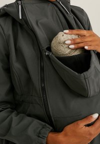 Donkergrijze hooded jacket met een ritszak, waarin een gebreide muts is opgeborgen; heeft elastische manchetten en een textuurstof voor warmte.