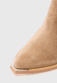 Bottine en daim beige avec un bout pointu et un embout en métal argenté sur une semelle en bois, le tout sur un fond clair.