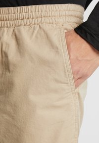 Shorts en coton beige avec une taille élastique, dotés de poches latérales et de détails de couture visibles. Texture lisse avec une coupe décontractée.