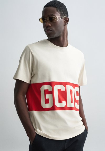 T-shirt en coton crème avec une large rayure horizontale rouge et le texte "GCDS" en blanc. Manches courtes et col rond. Texture lisse.