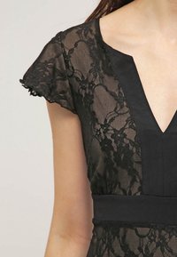 Robe en dentelle noire avec un décolleté en V profond et des manches volantées, dotée d'une taille noire unie et d'un motif de dentelle florale complexe.