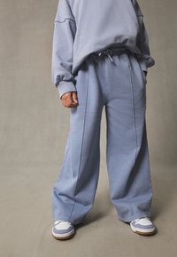 Pantalon de survêtement bleu clair, à jambes larges, avec une taille à cordon de serrage et une texture délavée. Associé à des baskets blanches et grises. Coupe décontractée et confortable.