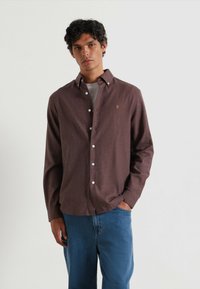 Camicia marrone a pulsante con una trama sottile e cuciture a contrasto, con bottoni bianchi e un logo ricamato sul petto.