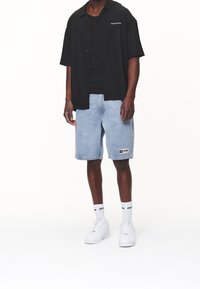 Maglietta nera a maniche corte sopra una canotta nera, abbinata a pantaloni corti in denim azzurro chiaro con una toppa color logo, scarpe da ginnastica bianche e calzini alla caviglia.
