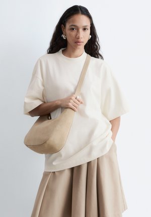 Giovane donna con capelli lunghi e scuri, indossa una maglietta beige a maniche corte larghe e una gonna, tiene una borsa a tracolla beige, rivolta verso avanti.