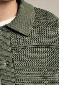 Cardigan verde lavorato a maglia con una superficie testurizzata, dotato di un colletto classico e dettagli a strisce orizzontali. Variazioni sottili nel motivo della maglia.
