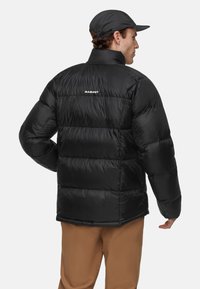 Homme portant une veste doudoune noire Mammut et une casquette noire, vu de dos, avec un pantalon marron sur un fond blanc uni.
