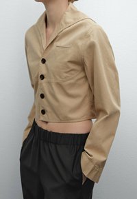 Person wearing a cropped beige button-up jacket with long sleeves and black elastic waist pants, hands in pockets, against plain background.