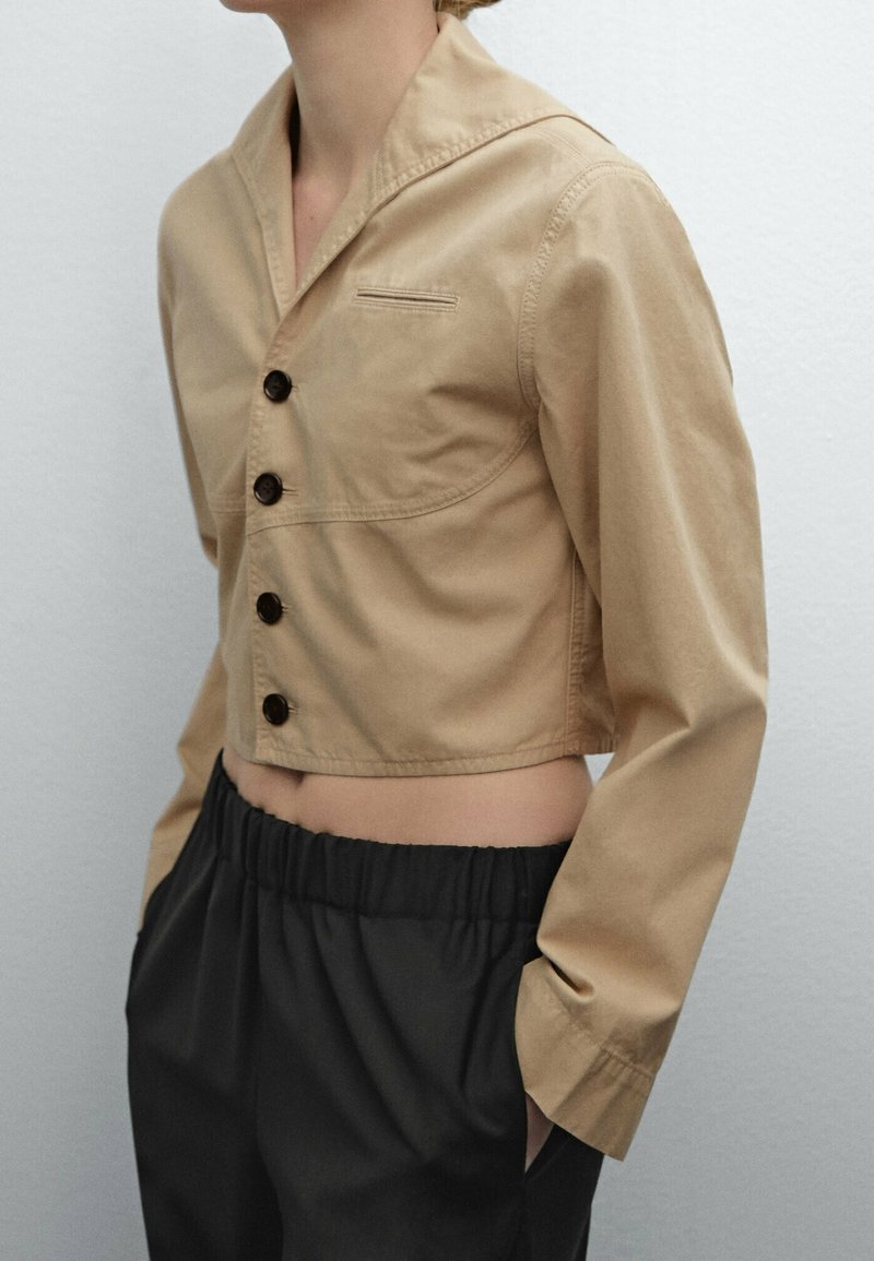 Person wearing a cropped beige button-up jacket with long sleeves and black elastic waist pants, hands in pockets, against plain background.