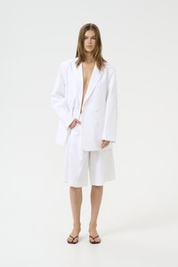 Blazer blanc à double boutonnage avec larges revers, associé à un short blanc au genou. Lignes simples et épurées, matériel minimal et sensation légère.