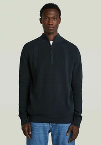 Donkergroen pullover sweater met een halve rits, geribbelde textuur, lange mouwen en een relaxte fit. Ggedragen over een wit t-shirt.