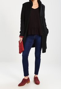 Donna in cappotto scuro e top nero, indossando jeans skinny blu e scarpe in suede rosse, che tiene una borsa a mano rossa abbinata nella mano sinistra.