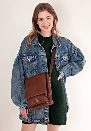Braune Leder-Crossbody-Tasche mit Klappe, getragen über einem dunkelgrünen Pullover und einer übergroßen Denimjacke, mit großen Taschen und Knopfdetails.