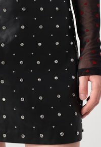 Jupe en cuir noire avec des clous argentés uniformément espacés, associée à une manche noire transparente avec des motifs de cœurs rouges et une main sur le côté.