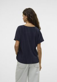 Marineblå plisseret top med korte ærmer og rund halsudskæring, parret med lyse, vide bukser. Glat tekstur og løst fit.