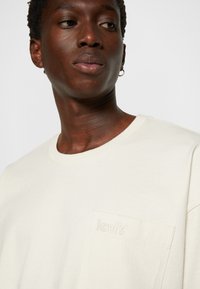 Cremefarvet t-shirt med korte ærmer lavet af bomuld, der har en lille lomme med et præget Levi's-logo på venstre side.