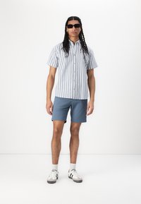 Chemise à manches courtes à rayures bleues claires et blanches, assortie à un short bleu. Les accessoires incluent des lunettes de soleil noires et des baskets.